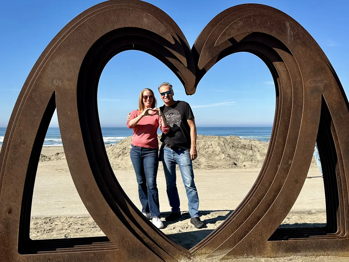a couple stands in front of a heart public art piece at sunset dunes park with the pacific ocean behind them